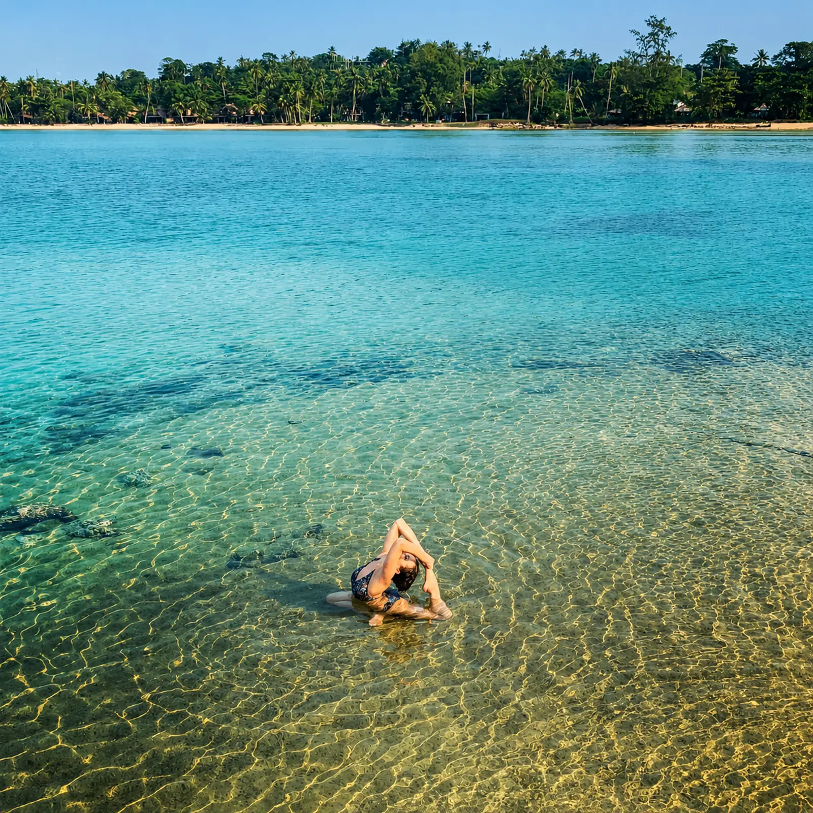 Woman practising yoga in the crystal-clear shallow water of Koh Mak, Thailand, with a palm-fringed beach in the background